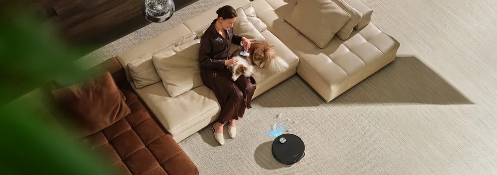 Dreame robot vacuum lifting pet hair from the carpet while a woman grooms her dog on the sofa