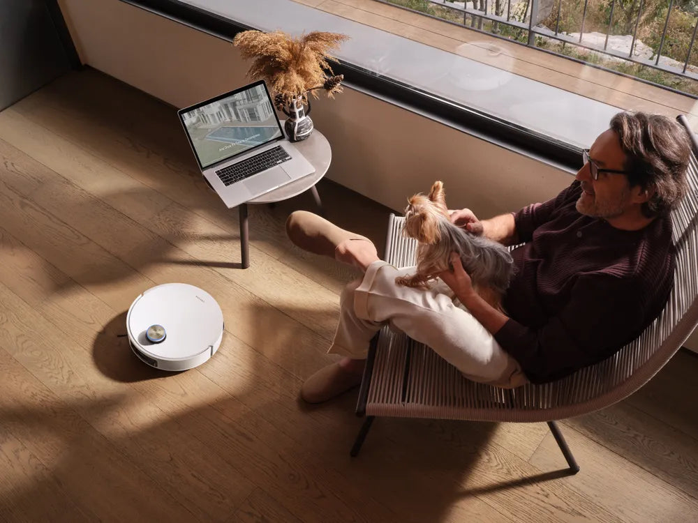 Man relaxes with his dog while a Dreame robot vacuum quietly cleans the apartment floor nearby