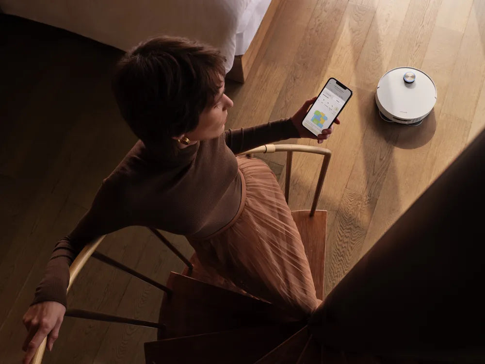 Woman sets no-go zones in the Dreame app while a robot vacuum cleans the floor nearby