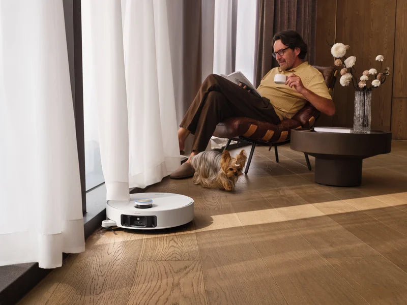Dreame robot vacuum cleaning hardwood floors beside a Yorkshire Terrier and its owner relaxing in an armchair.