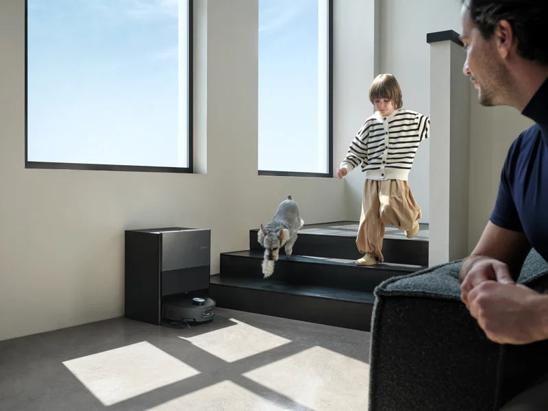 Dreame robot vacuum docked at its base station while a child and dog play on nearby stairs, father watching in the foreground.