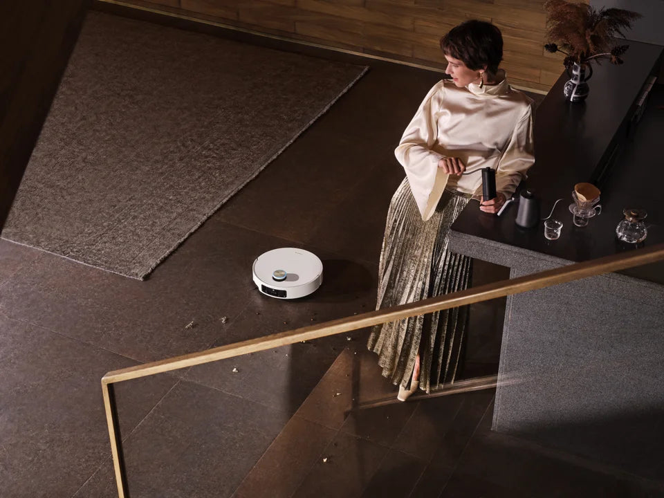 Dreame robot vacuum cleaning a dark tile floor with carpet and debris visible nearby, woman standing at a kitchen counter above.